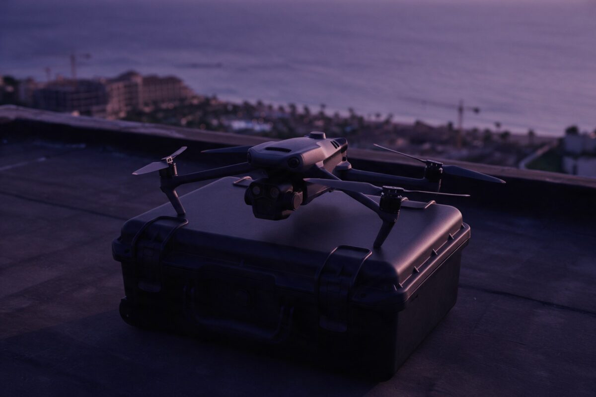 Akşamüstü bir çatıda sert çanta üzerinde duran profesyonel drone, arkada bulanık Akdeniz kıyısı, bir tatil tesisi ve vinçli şantiye