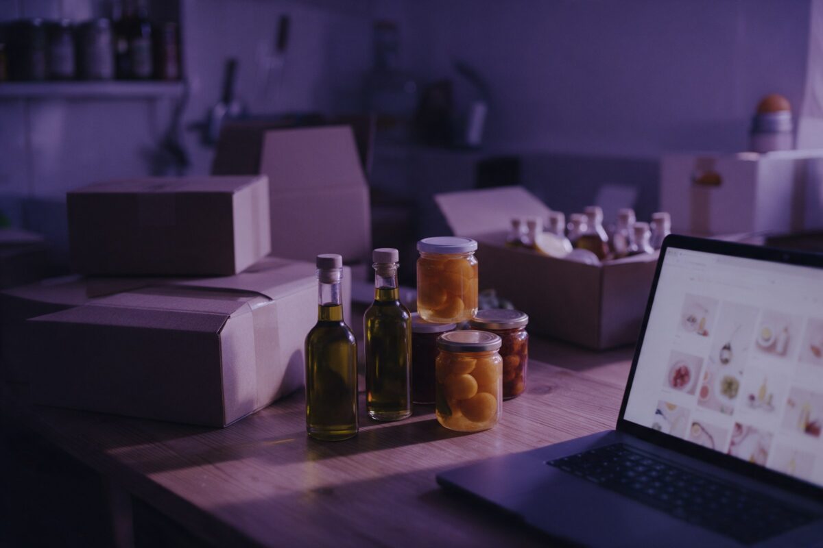 Küçük bir üretici paketleme odasında ahşap masada etiketsiz zeytinyağı ve reçel kavanozları, kraft kargo kutuları ve soyut çevrim içi mağaza ürün ızgarası gösteren dizüstü bilgisayar